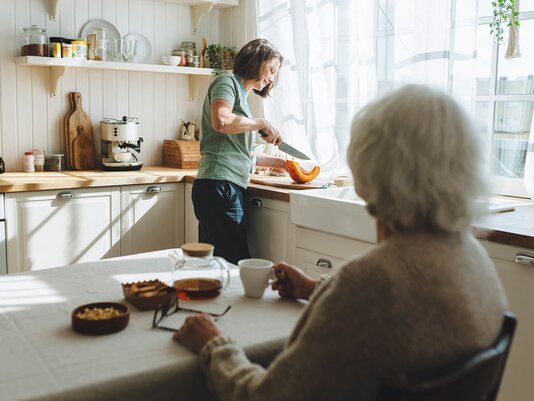 A Helper prepares a fresh meal in a sunlit kitchen while an older woman sits at the table with tea, waiting and watching.