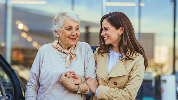 A senior woman and her Helper smile at each other while walking arm-in-arm outside a building. They look happy and relaxed, enjoying each other’s company. The mood is warm and cheerful.