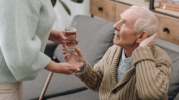 A senior man seated on a couch looks up while reaching for a glass of water and pills being handed to him by another person.