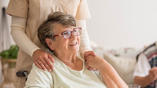 Herewith Helper standing behind a senior woman, hands on her shoulders, offering comfort and personal care.