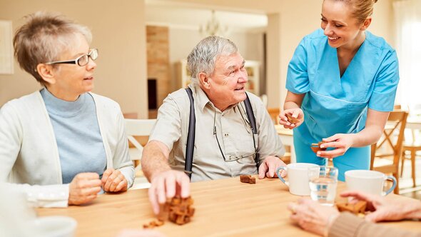 A group of older adults and a Helper share a playful moment around a table with wooden puzzle pieces. Everyone looks animated and connected—brain games and bonding rolled into one.