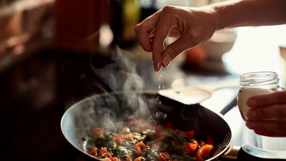 A close-up of a hand sprinkling salt over a skillet filled with steaming chopped vegetables on a stovetop.