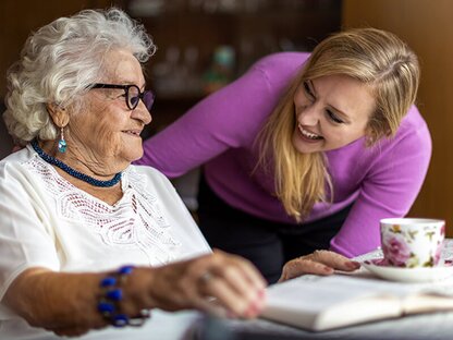 A Helper assists a senior woman with reading and daily engagement activities at home. Herewith provides flexible in-home senior care focused on dignity, independence, and meaningful connection.