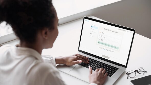 A person sits at a desk and uses a laptop to select scheduling options for help on the Herewith platform.