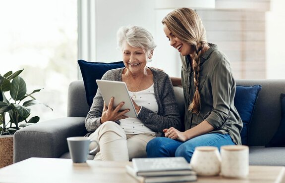 Helper and an older woman smile together on a sofa while looking at a tablet