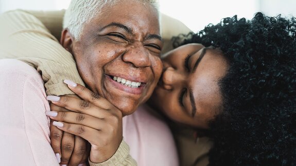 A woman hugging and kissing her mother on the cheek with joy and affection, both smiling and laughing.