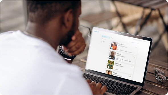 A person sits at a table using a laptop to view a list of Helper profiles on the Herewith platform.