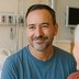 A man in a blue shirt sits beside someone in a hospital room and smiles.