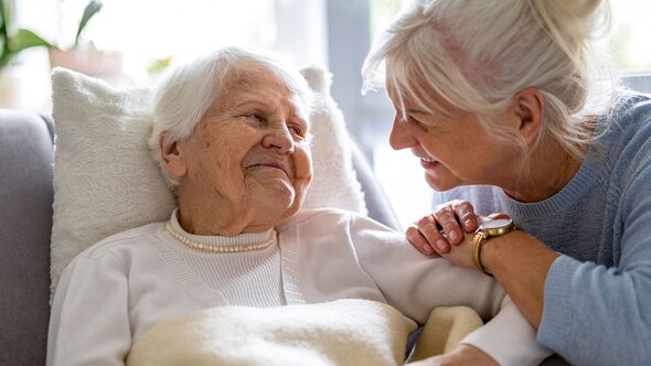 A woman leans in to speak gently with an older woman lying in bed, resting a hand on her arm.