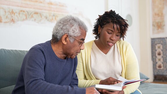 A Helper and a senior man sit together on a couch looking at a book, with the Helper reading aloud.