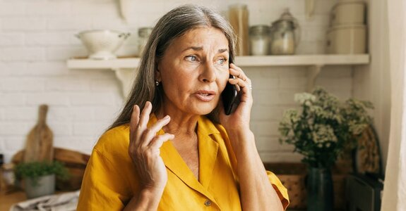 A woman stands in a kitchen holding a phone to her ear, gesturing with her hand as she speaks.