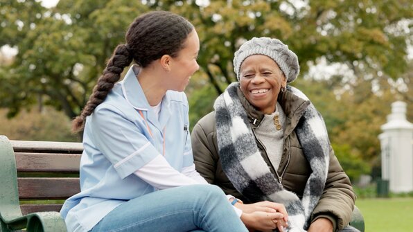 A senior woman sits on a bench in the park with a Helper. They're smiling and happy. 