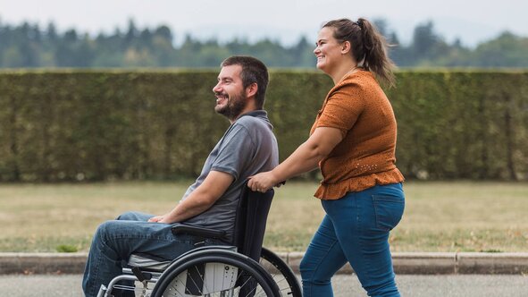 A Helper walks alongside a man in a wheelchair on a sunny path, both smiling.