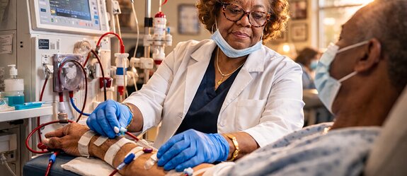 Doris Jones, a Black woman in a white coat and surgical mask, carefully connects a patient's arm to a dialysis machine in a treatment center.