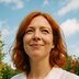 A woman with red hair looks ahead while standing outdoors with trees and sky in the background.