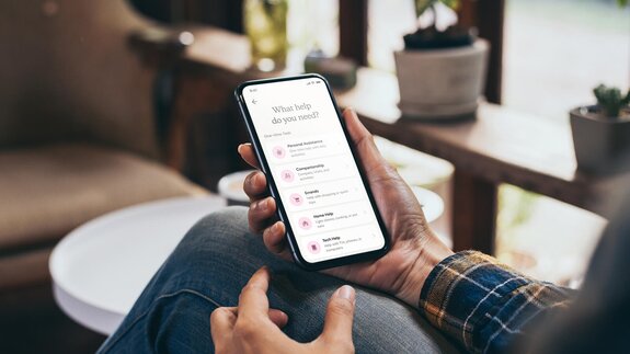 A person holds a smartphone showing various categories of help including personal assistance, companionship, and errands.