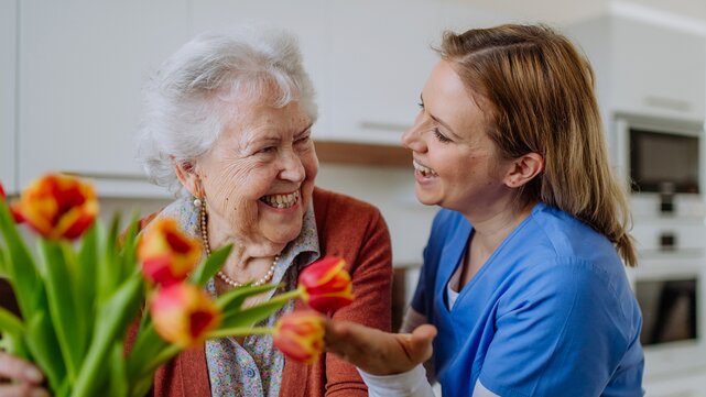 A helper shows off some pretty red flowers to a senior. They're smiling in the kitchen. 