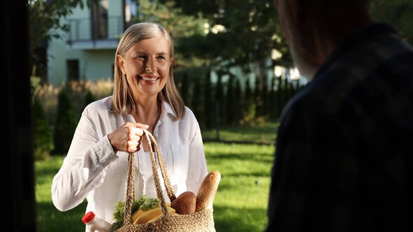A Helper delivers a basket of fresh groceries to an older man at his front door.