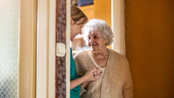 A senior woman stands in a doorway, smiling warmly while holding hands with a Helper. They share a tender, joyful moment, suggesting trust and companionship.
