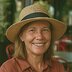A woman in a brown shirt and straw hat smiles at the camera while sitting outdoors.