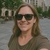 A woman wearing sunglasses takes a selfie in a city plaza with people in the background.