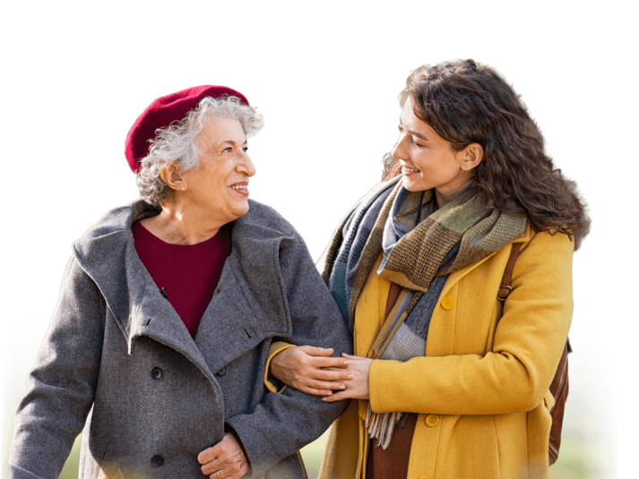 A senior and a helper walk arm in arm smiling and laughing together. The senior has short curly hair, a red beret and gray jacket on. The Helper has a scarf and a long yellow coat. 