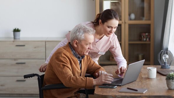 Herewith Helper assisting a senior man in a wheelchair with a laptop, supporting technology and daily tasks at home.