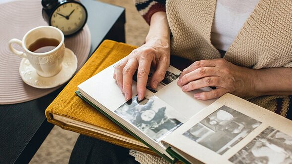 A person sitting at a table turns pages in a photo album filled with black-and-white pictures, with a cup of tea and a clock nearby.