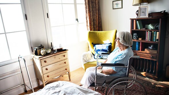 An older woman sits quietly in a wheelchair, gazing out a sunny window in a cozy room. The mood is peaceful but slightly pensive, with soft morning light filling the space.