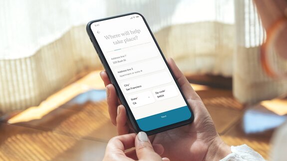 Close-up of a phone screen showing a form to enter the service location, held by someone indoors near soft window light.