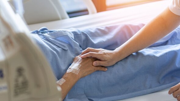 A hand rests on the hand of a patient in a hospital bed with a visible IV site on their arm.