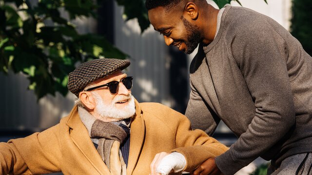 A Helper supports an older man wearing a coat and scarf as they share a moment outside.