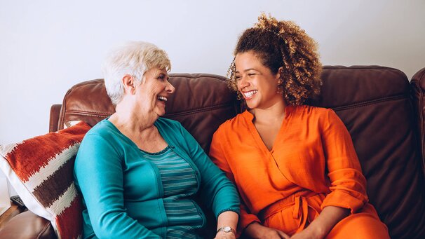 A Helper dressed in orange, sits and chats happily with a Senior on the sofa. 