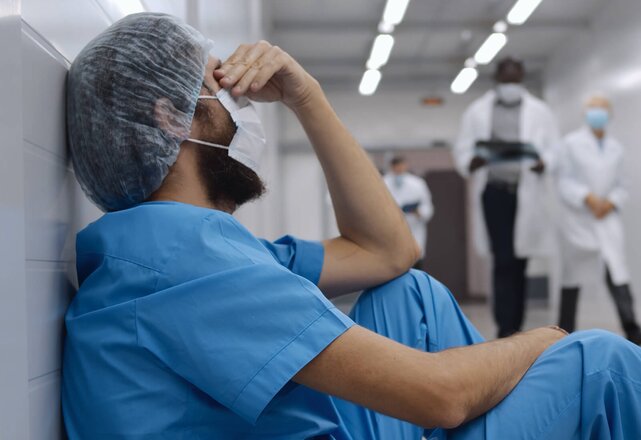 Overwhelmed Healthcare Professional sits on the floor in the hospital. They're wearing scrubs and PPE and holding their head