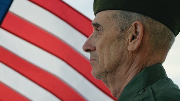 A senior man in military uniform stands in profile with a serious expression. An American flag waves prominently in the background, symbolizing patriotism and service.