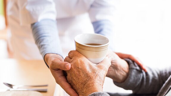 A Helper hands a cup to an older adult who is reaching out with both hands to receive it.