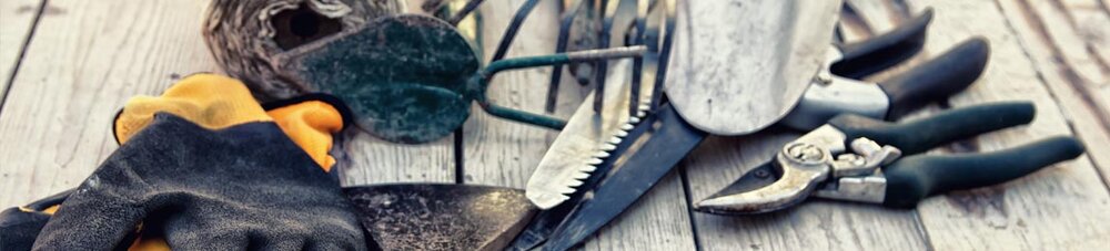 Verschiedene Gartenwerkzeuge, darunter Handschuhe, Axt, Schaufel, Rechen, Astschere und Schnur, auf Holzboden ausgelegt.