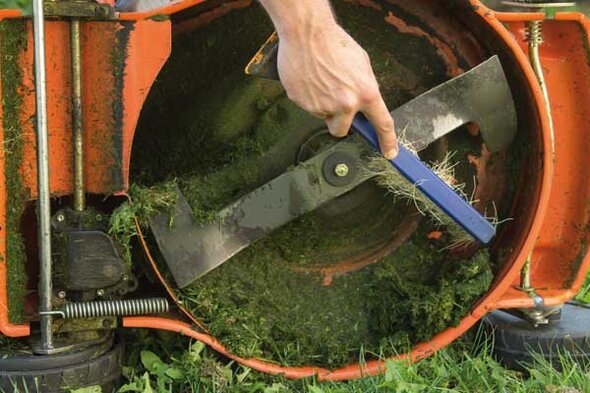 Hand reinigt mit einer Bürste das Messergehäuse eines auf den Kopf gedrehten Rasenmähers von Grasresten.