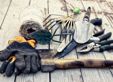 Verschiedene Gartenwerkzeuge, darunter Handschuhe, Axt, Schaufel, Rechen, Astschere und Schnur, auf Holzboden ausgelegt.