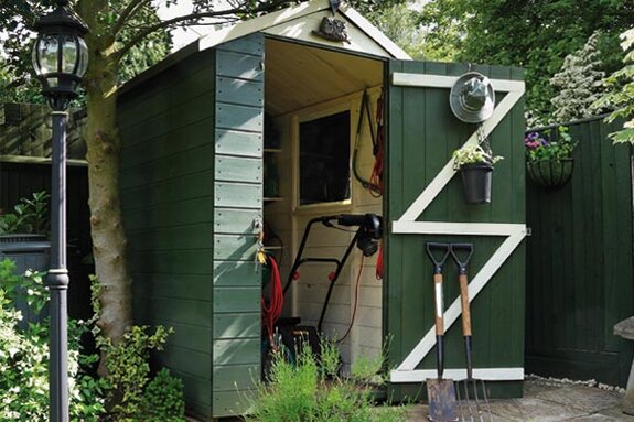 Geöffnetes grünes Gartenhaus mit Werkzeugen, Rasenmäher im Inneren und Schaufeln an der Tür, umgeben von Pflanzen.