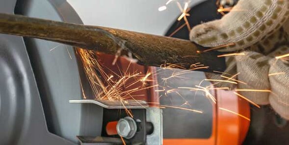 Schleifvorgang mit Funkenflug: Eine Hand mit Arbeitshandschuh hält ein Metallmesser an eine rotierende Schleifscheibe.