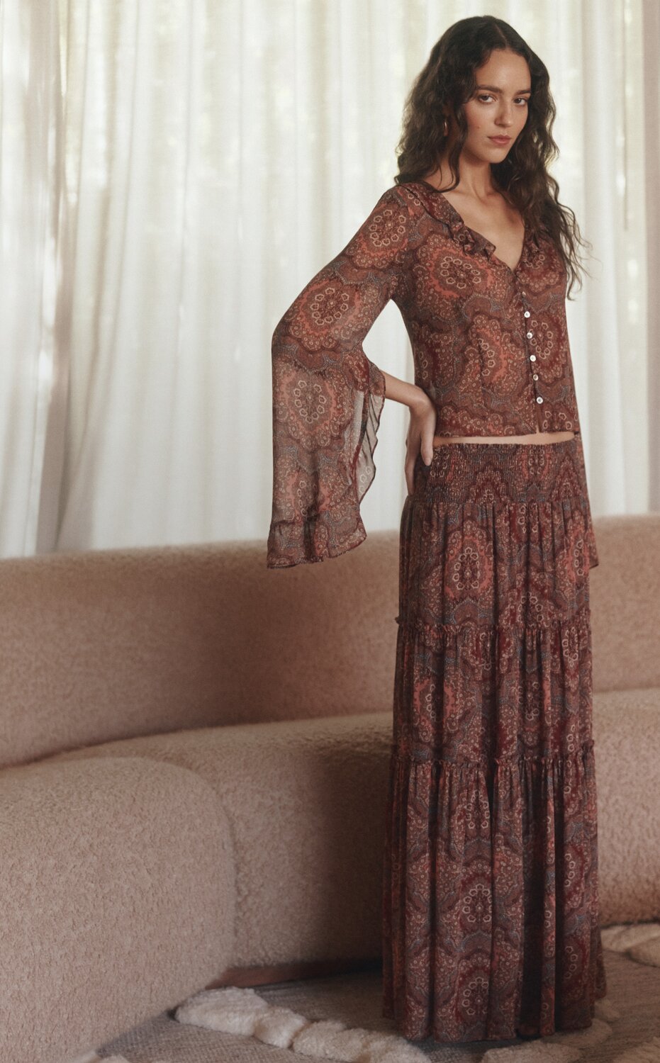 A woman posing in front of a sofa wearing a red paisley printed silk blouse with a matching red paisley printed silk skirt