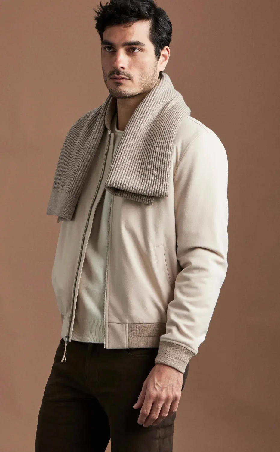 A man standing wearing a cream bomber jacket and sand scarf