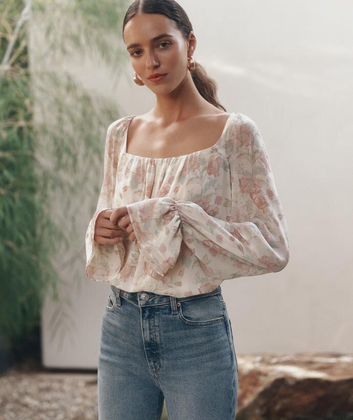 A woman wearing a beautiful silk long sleeve, belled cuff blouse in a beige, pink and light mint colored floral pattern paired with medium blue wash denim jeans in a garden setting.
