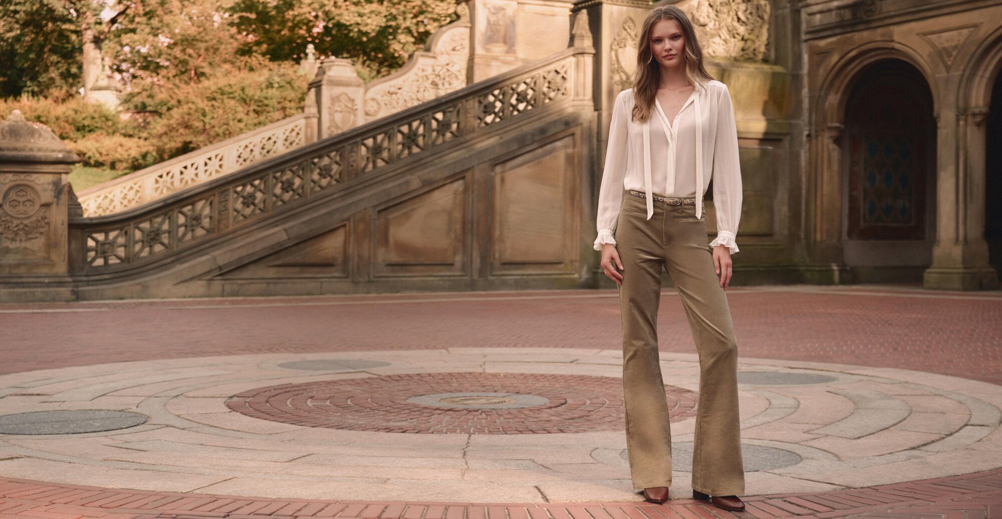Woman wearing a white long sleeve blouse with olive green corduroy pants, a slim snake printed belt, and brown boots