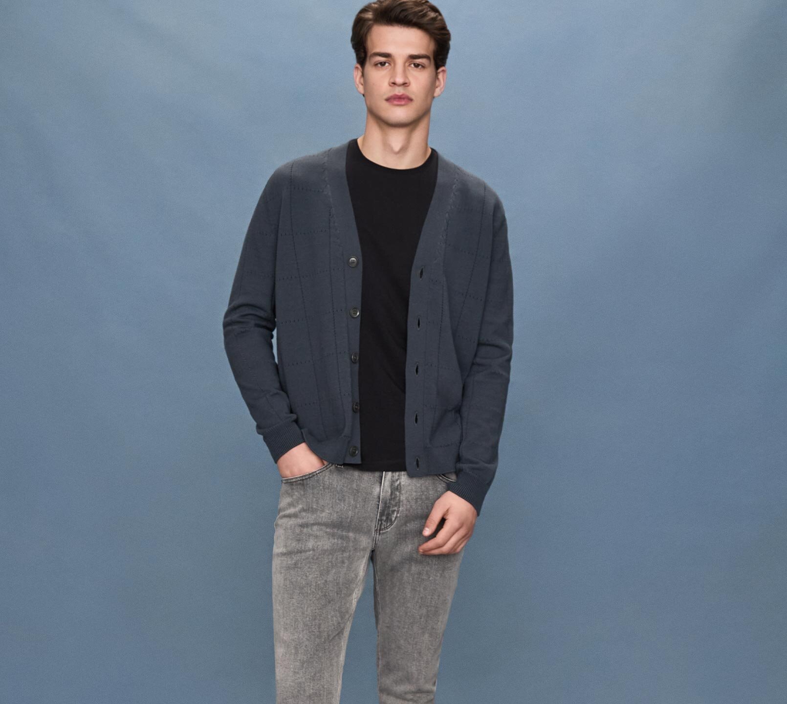 Man standing wearing a grey v-neck sweater over a grey button up shirt, dark wash slim fit jeans and black sneakers