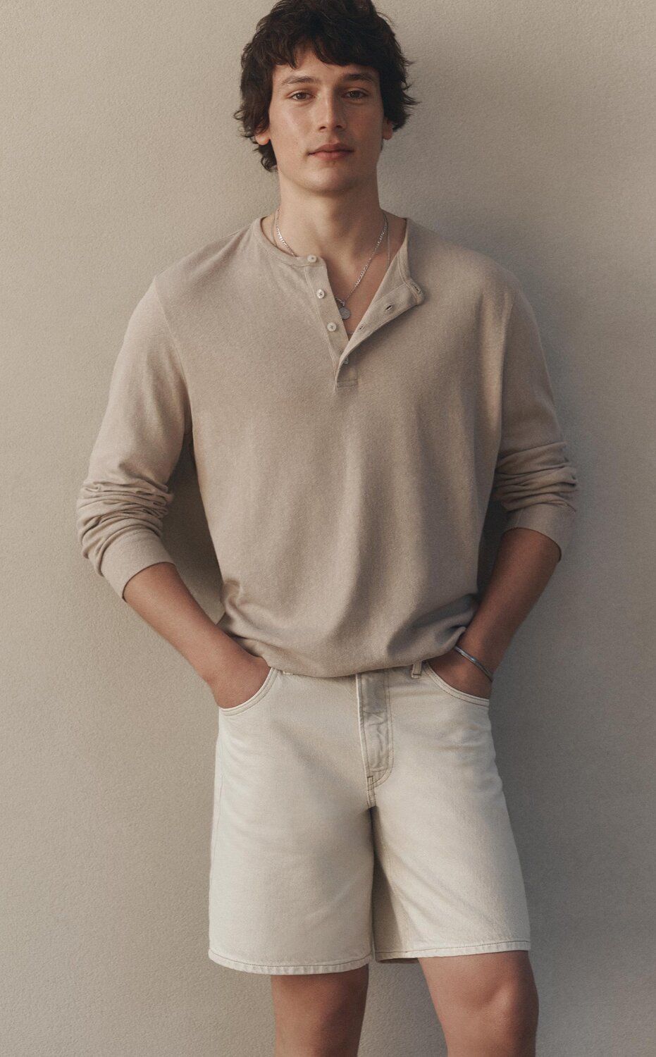 Man wearing a long-sleeve henley shirt and light shorts, standing against a neutral wall.