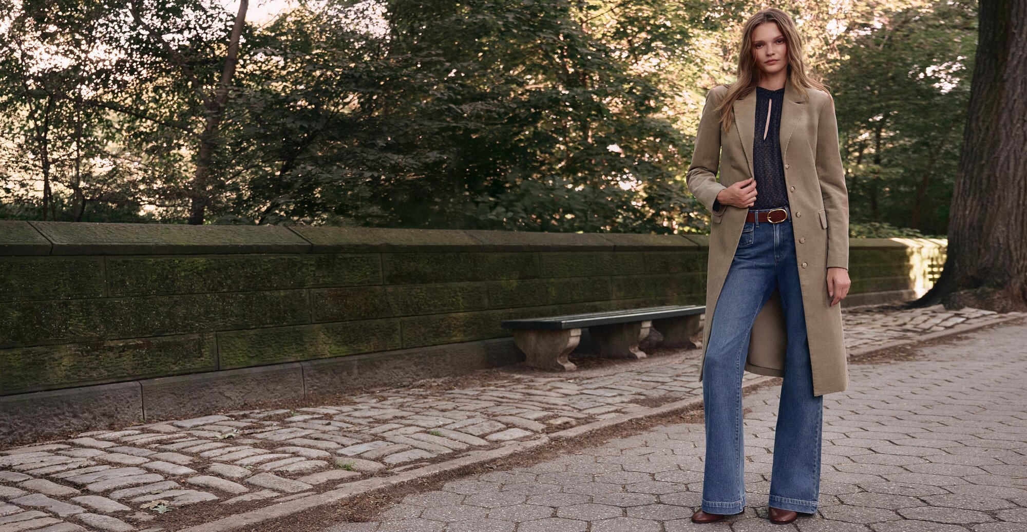 Woman wearing a long olive green corduroy coat over a blue blouse, wide leg jeans, a brown belt and brown boots