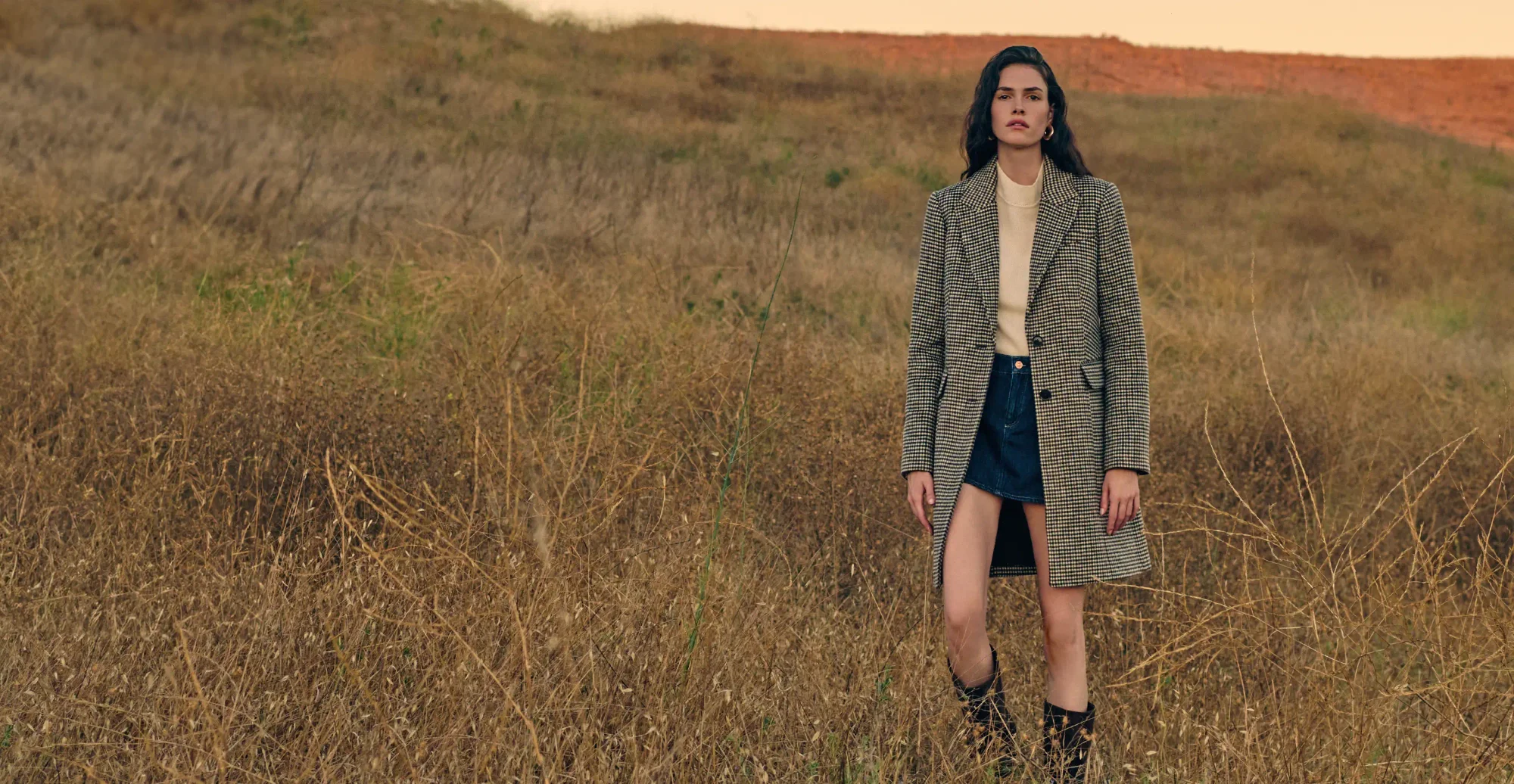 Woman standing in a field wearing a long houndstooth coat, an ivory sweater, denim mini skirt, and tall black boots