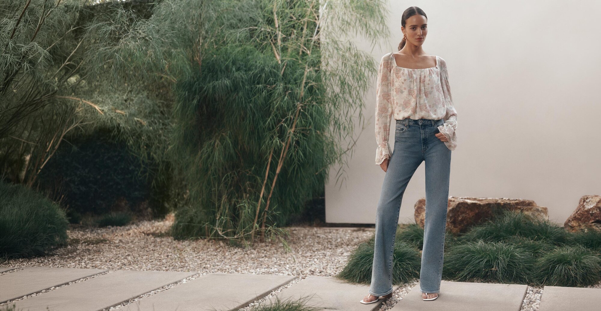 Woman wearing a long-sleeve floral blouse and light-wash high-rise bootcut jeans standing outdoors against a cream wall and green foliage.